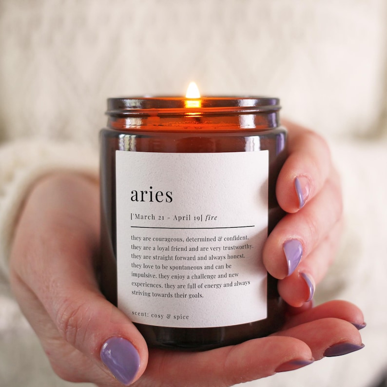 May include: A brown glass candle with a white label that reads "aries [March 21 - April 19] fire" and a description of the astrological sign aries. The candle is lit and the scent is described as "cosy & spice".