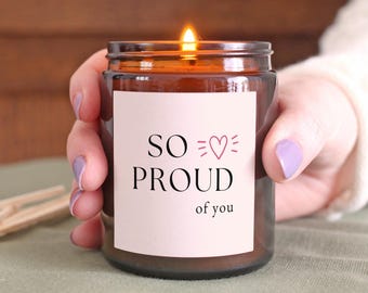 May include: A burning candle in a brown glass jar. The jar features a white label with the text "SO PROUD of you" in black, and a pink heart. The candle is held by a person.