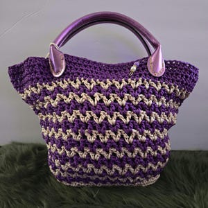 May include: A handcrafted crochet tote bag in a vibrant purple and beige chevron pattern. The bag features a sturdy design with a rounded shape, a purple leather handle, and a decorative bead accent. This stylish bag is perfect for carrying essentials.