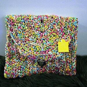 May include: A colorful, hand-woven clutch with a flap closure and a metal clasp. The bag is made of interwoven strips of fabric in various colors, including pink, yellow, blue, and green. A yellow tag hangs from the front.