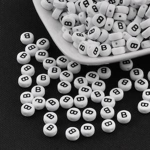 Könnte beinhalten: Weiße Kunststoffbuchstabenperlen mit dem Buchstaben "B" in Schwarz gedruckt. Die Perlen sind auf einer schwarzen Oberfläche verstreut.