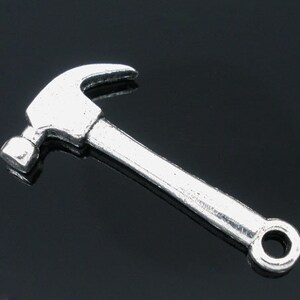 May include: A silver-colored hammer charm with a curved claw and a flat striking face. The hammer has a long handle and a small hole at the end. The charm is likely made of metal and is set against a dark background.
