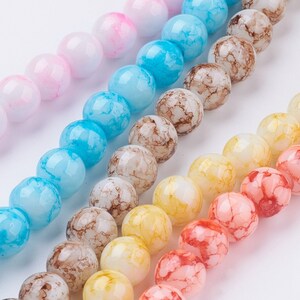 Può includere: Assortimento di perline di vetro colorate con effetto marmoreo. Le perline sono rotonde e hanno una finitura traslucida. I colori includono rosa, blu, giallo e arancione.