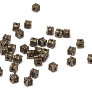 Könnte beinhalten: Eine Sammlung antiker, goldfarbener Metall-Spacer-Perlen mit filigranen Designs. Die Perlen sind quadratisch mit einem Loch in der Mitte.