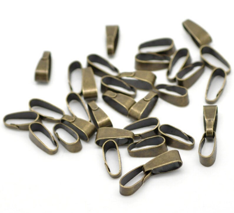 May include: A collection of antique bronze-colored pendant bails, scattered on a white surface. The bails have a loop shape with a small, rectangular top for attaching to a chain or cord. Ideal for jewelry making.