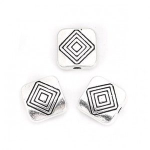 Puede incluir: Tres cuentas cuadradas plateadas con un diseño geométrico negro. Cada cuenta tiene un agujero en el centro para enhebrar.