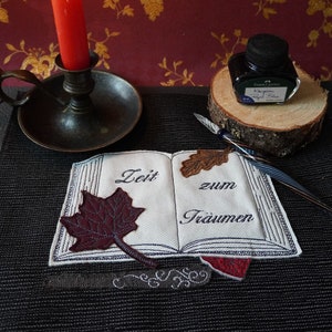 Op de afbeelding: Een boek met de tekst "Zeit zum Träumen" geborduurd op de omslag, een brandende kaars, een inktpot en een kalligrafiepen op een zwart oppervlak.