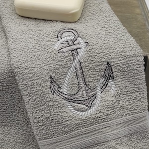 Könnte beinhalten: Zwei graue Handtücher mit einem gestickten Anker-Design. Ein weißer Stück Seife liegt auf den Handtüchern.