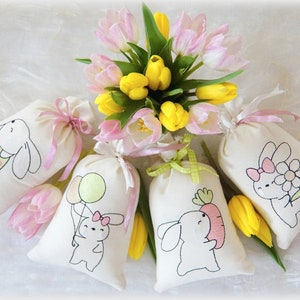 Könnte beinhalten: Vier weiße Stoffgeschenktüten mit Hasendesigns. Jede Tasche hat ein anderes Hasendesign, darunter ein Hase, der eine Karotte hält, ein Hase, der Ballons hält, ein Hase, der eine Blume hält, und ein Hase, der ein Osterei hält. Die Taschen sind mit rosa und grünen Bändern gebunden. Ein Strauß rosa und gelber Tulpen befindet sich in der Mitte des Bildes.