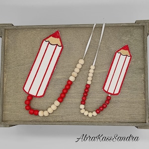 Puede incluir: Dos adornos en forma de lápiz rojo y blanco con una cadena de cuentas rojas y de madera natural. Los adornos cuelgan de una bandeja de madera con el texto "AbraKassSandra" en la parte inferior.