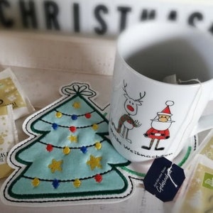 May include: A white ceramic mug with a Christmas design featuring a reindeer and Santa Claus. The mug is sitting on a coaster with an embroidered Christmas tree design. There are also tea bags in the background.