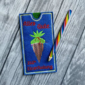 May include: A blue felt card with the text "Alles Gute zur Einschulung" embroidered on it. The card features a green and red embroidered carrot with a green stem and a brown string tied around it. A rainbow colored pencil is lying next to the card.