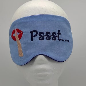 Op de afbeelding: Een lichtblauw slaapmasker met de geborduurde tekst "Pssst..." en een rood lippenontwerp. Het slaapmasker is ontworpen om licht te blokkeren en ontspanning te bevorderen. Het masker is gemaakt van zachte stof.