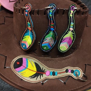 Puede incluir: Cuatro llaveros de plumas bordadas de colores con un diseño de cuentas. Los llaveros están unidos a un sombrero de vaquero marrón.