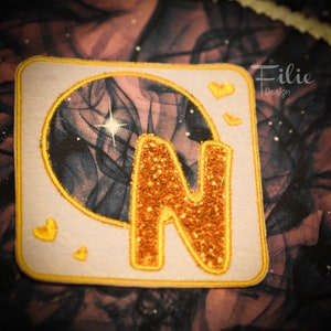 May include: A light purple square patch with a yellow border and a gold glitter letter N inside. The letter N is surrounded by a circle with a black and white galaxy design. There are three small yellow hearts in the top right corner of the patch.