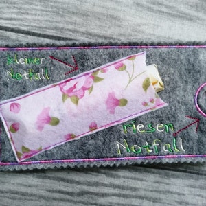 Puede incluir: Una bolsa de fieltro gris con un bolsillo de tela floral rosa. La bolsa tiene texto bordado que dice "kleiner Notfall" y "riesen Notfall" con flechas apuntando a cada uno.