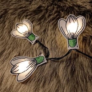 May include: A string of decorative lights featuring three white, flower-shaped fabric cutouts with green accents and black stitching. The lights are illuminated, creating a warm glow. The string is on a brown, furry surface.