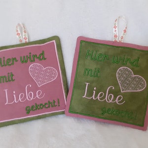 May include: Two green and pink pot holders with a heart and the text "Hier wird mit Liebe gekocht!"  in green embroidery.