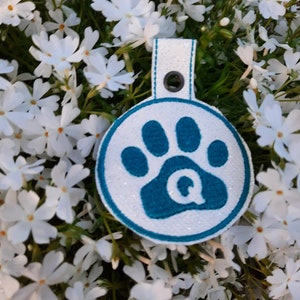Op de afbeelding: Een wit en turquoiseblauw hondenpenning met een pootprint en de letter Q in het midden. De penning is omringd door witte bloemen.