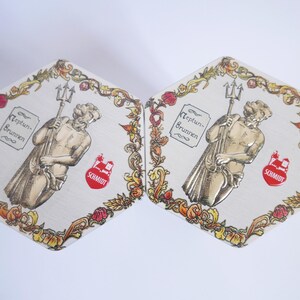 May include: Two hexagonal coasters with a white background and a gold and red floral border. Each coaster features a drawing of a man holding a trident, with the text "Neptun-Brunnen" and "SCHMIDT" in red.