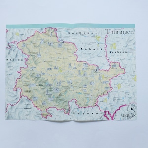 May include: A folded map of Thuringia, Germany, with a light blue border. The map features a detailed depiction of the region, with pink borders outlining the area and various place names.