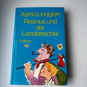 Könnte beinhalten: Ein gebundenes Buch mit dem Titel "Astrid Lindgren Rasmus und der Landstreicher" in deutscher Sprache. Das Cover ist hellblau mit gelbem Text und einer farbenfrohen Illustration von zwei Figuren. Ein Mann mit Pfeife und ein Junge sind abgebildet.