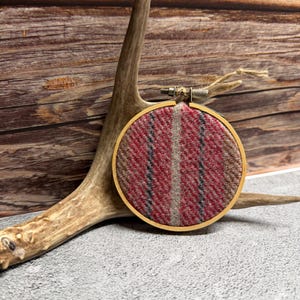 May include: A round embroidery hoop with a red, gray, and brown plaid fabric design. The hoop is made of wood and is held in place by a metal clasp. The background features a deer antler and a wooden surface.
