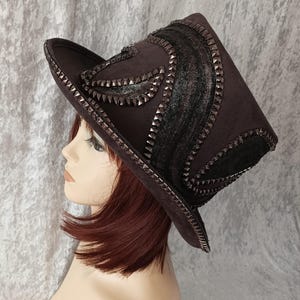 Puede incluir: Un sombrero de copa negro con un ala ancha y un diseño decorativo de tela negra y tachuelas plateadas. El sombrero está inclinado hacia un lado y lo lleva un maniquí.