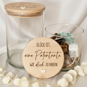 Pode incluir: Dois frascos de vidro transparente com tampas de madeira e uma tampa de madeira com o texto "Glück ist, eine Patentante wie dich zu haben!" Uma grinalda floral branca está drapeada sobre a superfície branca.