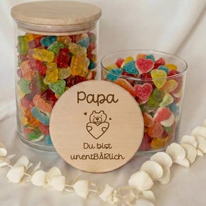 Könnte beinhalten: Zwei Gläser gefüllt mit bunten Gummibonbons, eines groß und eines klein, neben einer Holzscheibe mit dem Wort "Papa" und einer Bärenillustration. Die Süßigkeiten haben verschiedene Formen und Farben.