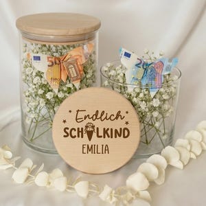 Può includere: Due barattoli in vetro trasparente con coperchi in legno chiaro, riempiti con fiori bianchi di gipsofila e farfalle origami realizzate con banconote in euro. Un coperchio circolare in legno riporta inciso il testo Endlich SCHULKIND EMILIA.