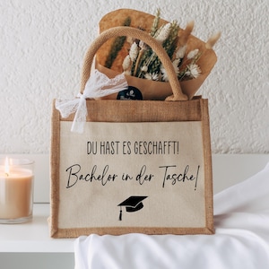 Op de afbeelding: Een afstudeercadeauset met een jute draagtas met de tekst "DU HAST ES GESCHAFFT! Bachelor in der Tasche!" en een afstudeerpet-icoon. De tas bevat gedroogde bloemen en staat naast een brandende kaars.