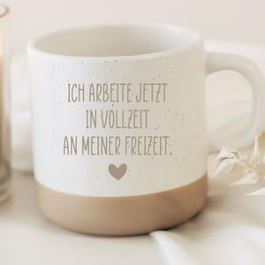 Könnte beinhalten: Ein weißer Keramikbecher mit gesprenkelter Oberfläche und beigem Boden. Der Becher trägt den grauen Aufdruck "ICH ARBEITE JETZT IN VOLLZEIT AN MEINER FREIZEIT." und ein kleines, mittig darunter platziertes graues Herzsymbol.
