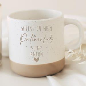 Könnte beinhalten: Eine weiße Keramik-Tasse mit gesprenkeltem Design und beigefarbenem Boden. Auf der Tasse steht der Text "WILLST DU MEIN Patenonkel SEIN? ANTON" und ein Herzsymbol. Eine brennende Kerze und weißer Stoff sind im Hintergrund.