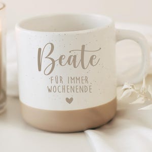 Könnte beinhalten: Eine weiße Keramik-Tasse mit gesprenkelter Oberfläche und beigem Boden. Die Tasse ist mit dem Namen Beate in Schreibschrift bedruckt, gefolgt von dem Text FÜR IMMER WOCHENENDE und einem kleinen Herz-Symbol in Dunkelgrau. Die Tasse hat einen Henkel.
