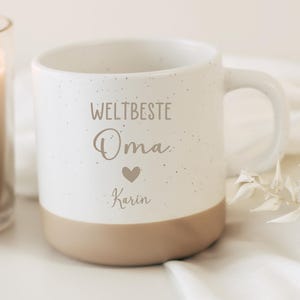 Op de afbeelding: Een witte keramische mok met een gespikkelde afwerking en een beige basis. De voorzijde bevat de tekst "WELTBESTE Oma" in een mix van lettertypes, een klein hartje en de naam "Karin" gedrukt in een sierlijk cursief lettertype.