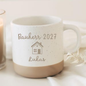Könnte beinhalten: Weißer Keramikbecher mit gesprenkeltem Design und beigefarbenem Boden. Der Becher trägt den Text "Bauherr 2027" über einem Haus-Symbol und dem Namen "Lukas". Im Hintergrund ist eine brennende Kerze zu sehen.