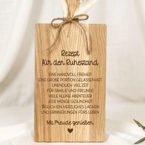 Peut inclure: Une planche à découper rectangulaire en bois avec une poignée percée d'un trou. La planche est gravée avec du texte en allemand et un petit cœur. Un nœud en ficelle brune avec des petites fleurs blanches séchées orne la poignée.