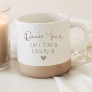 Pode incluir: Uma caneca de cerâmica branca com acabamento salpicado e base bege. A frente apresenta o texto "Danke Mama, FÜR ALL DIE GROBEN & KLEINEN DINGE!" numa fonte cinzenta escura manuscrita e sem serifa, com um pequeno coração centrado abaixo.