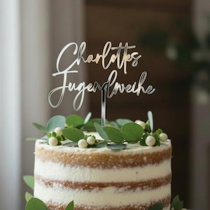 Op de afbeelding: Een ronde, gelaagde taart met witte frosting en een zilveren taarttopper met de tekst "Charlottes Jugendweihe". De taart is versierd met groene bladeren en witte bessen en staat op een houten taartstandaard.