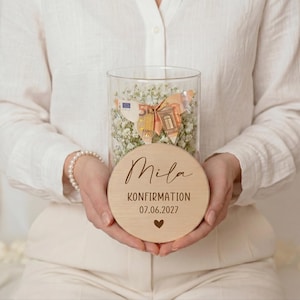 May include: A clear glass jar filled with white flowers and money, topped with a wooden disc. The disc is engraved with the name "Mila", the word "KONFIRMATION", the date "07.06.2027", and a heart symbol. The jar is held by a person.