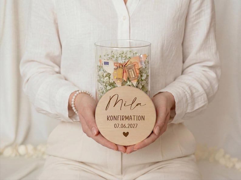 May include: A clear glass jar filled with white flowers and money, topped with a wooden disc. The disc is engraved with the name "Mila", the word "KONFIRMATION", the date "07.06.2027", and a heart symbol. The jar is held by a person.
