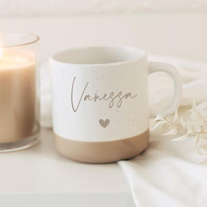May include: A white ceramic mug with the name "Vanessa" in a cursive font and a small heart. The bottom of the mug is a tan color. A lit candle in a glass jar is to the left of the mug.