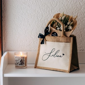 May include: A brown burlap tote bag with a white fabric panel that says "Selina" in black script with a small black heart. The bag is filled with a bouquet of dried flowers and a black ribbon.