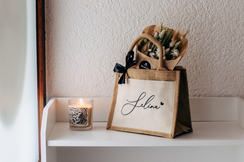 May include: A brown burlap tote bag with a white fabric panel that says "Selina" in black script with a small black heart. The bag is filled with a bouquet of dried flowers and a black ribbon.