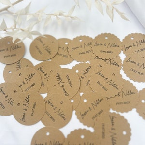 May include: A collection of brown paper circle tags with the text "Laura & Niklas" and the date "19.07.2025" printed on them. The tags have a hole punched in the top and some have scalloped edges.
