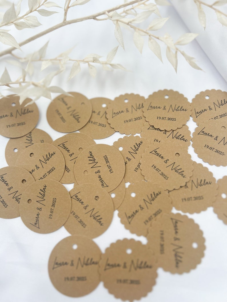 May include: A collection of brown paper circle tags with the text "Laura & Niklas" and the date "19.07.2025" printed on them. The tags have a hole punched in the top and some have scalloped edges.