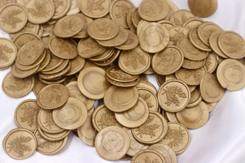 Op de afbeelding: Een stapel gouden waszegels met een bladerenontwerp. De zegels zijn rond en hebben een gladde, glanzende afwerking.