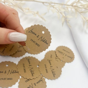 May include: Brown paper circle tags with scalloped edges and a hole punched in the top. The tags have the text "Laura & Niklas" and the date "19.07.2025" printed in black.