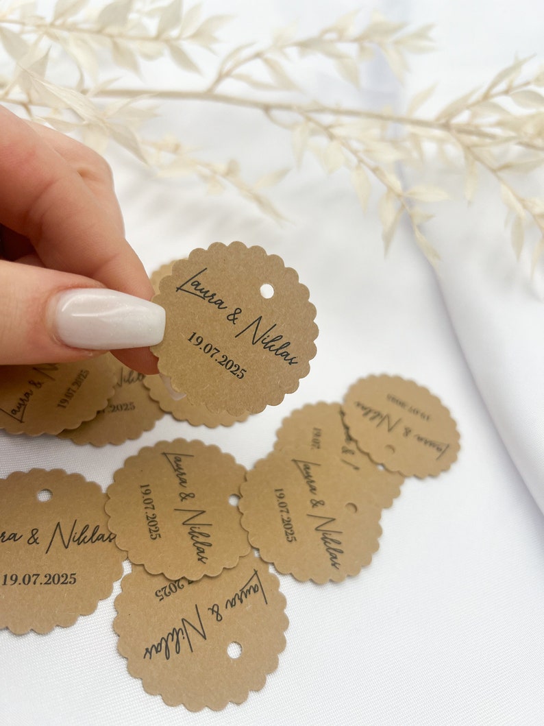 May include: Brown paper circle tags with scalloped edges and a hole punched in the top. The tags have the text "Laura & Niklas" and the date "19.07.2025" printed in black.